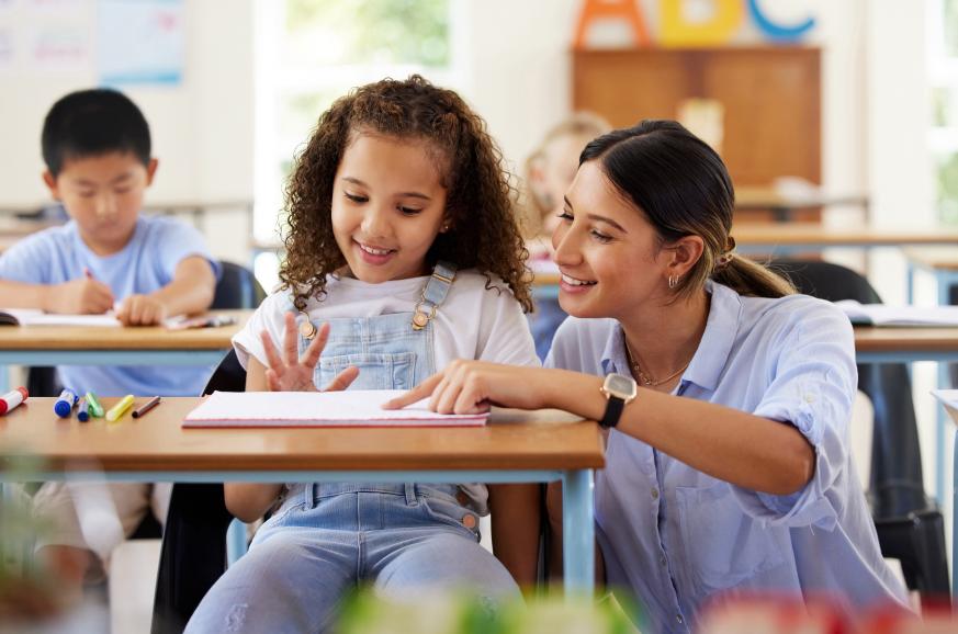 Teacher kneeling down and helping a female student who is sitting in her desk learning 