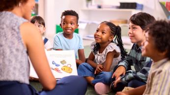Students sitting down, smiling at teacher who is reading.