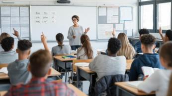 Teacher standing in front of a full room of students who are sitting at their desks and showing engagement in the lesson by raising their hands