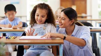 Teacher kneeling down and helping a female student who is sitting in her desk learning 