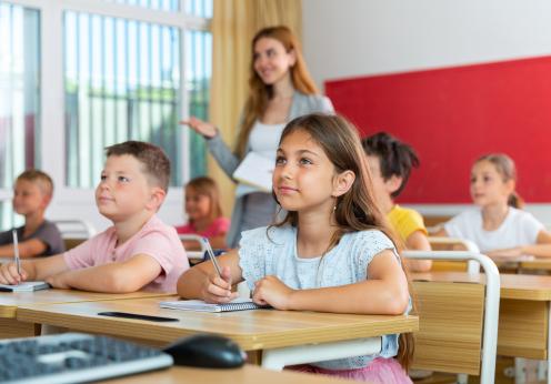 Student sitting at desk in classroom with teacher present.