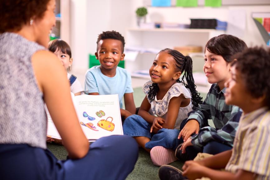 Students sitting down, smiling at teacher who is reading.