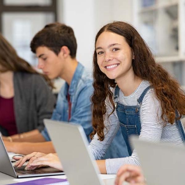 girl looking at camera while at table with other students with laptops